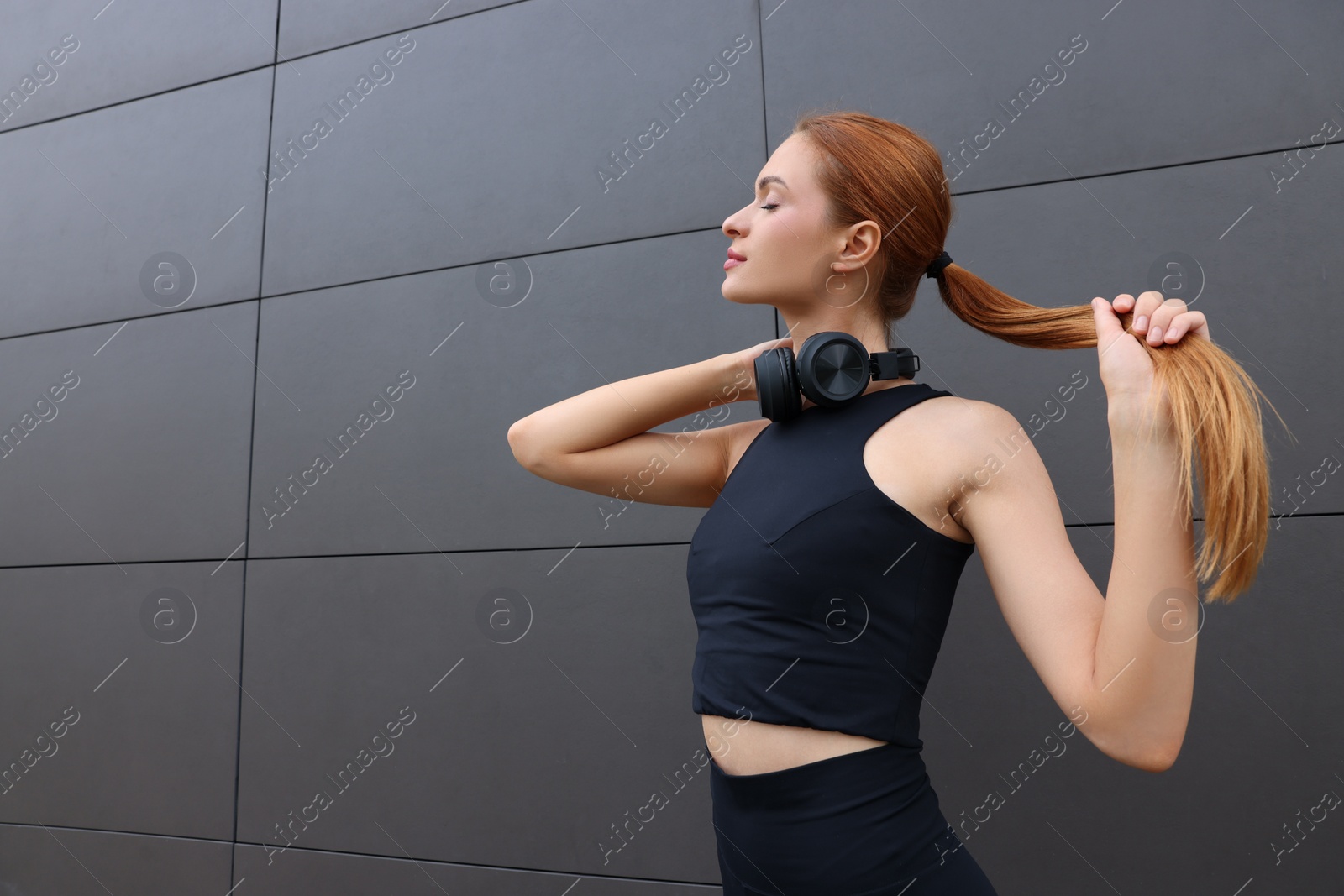 Photo of Beautiful woman in stylish gym clothes with headphones posing near dark grey wall on street, space for text