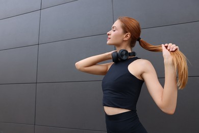 Beautiful woman in stylish gym clothes with headphones posing near dark grey wall on street, space for text