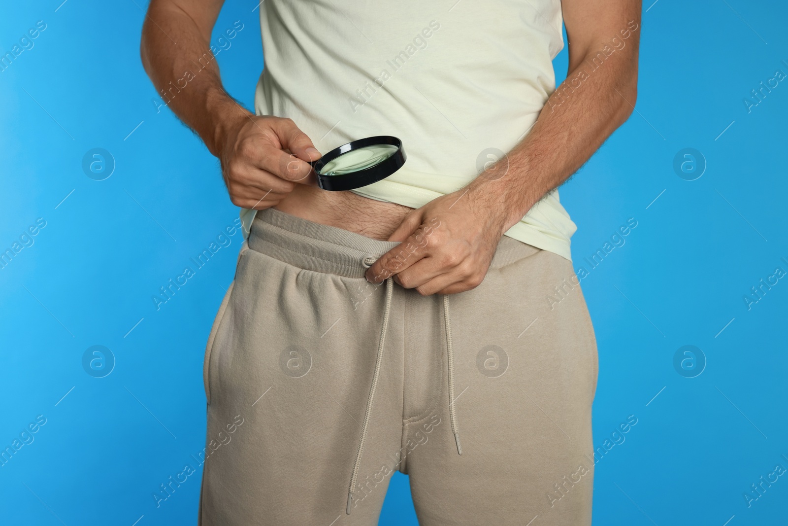 Photo of Man examining intimate area with magnifying glass on light blue background, closeup. Genital herpes