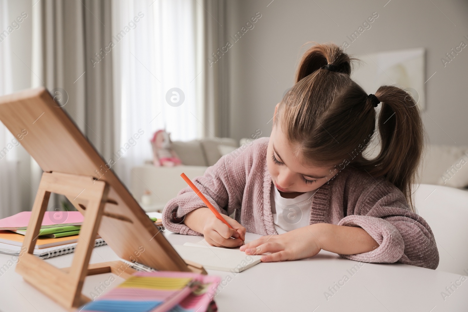 Photo of Adorable little girl doing homework with tablet at table indoors