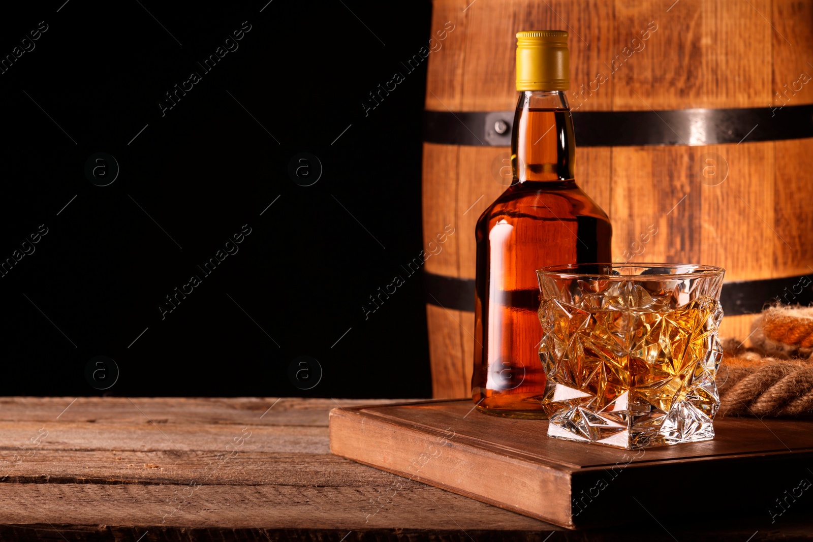 Photo of Whiskey with ice cubes in glass, bottle and barrel on wooden table against black background, space for text