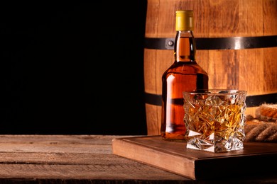 Photo of Whiskey with ice cubes in glass, bottle and barrel on wooden table against black background, space for text