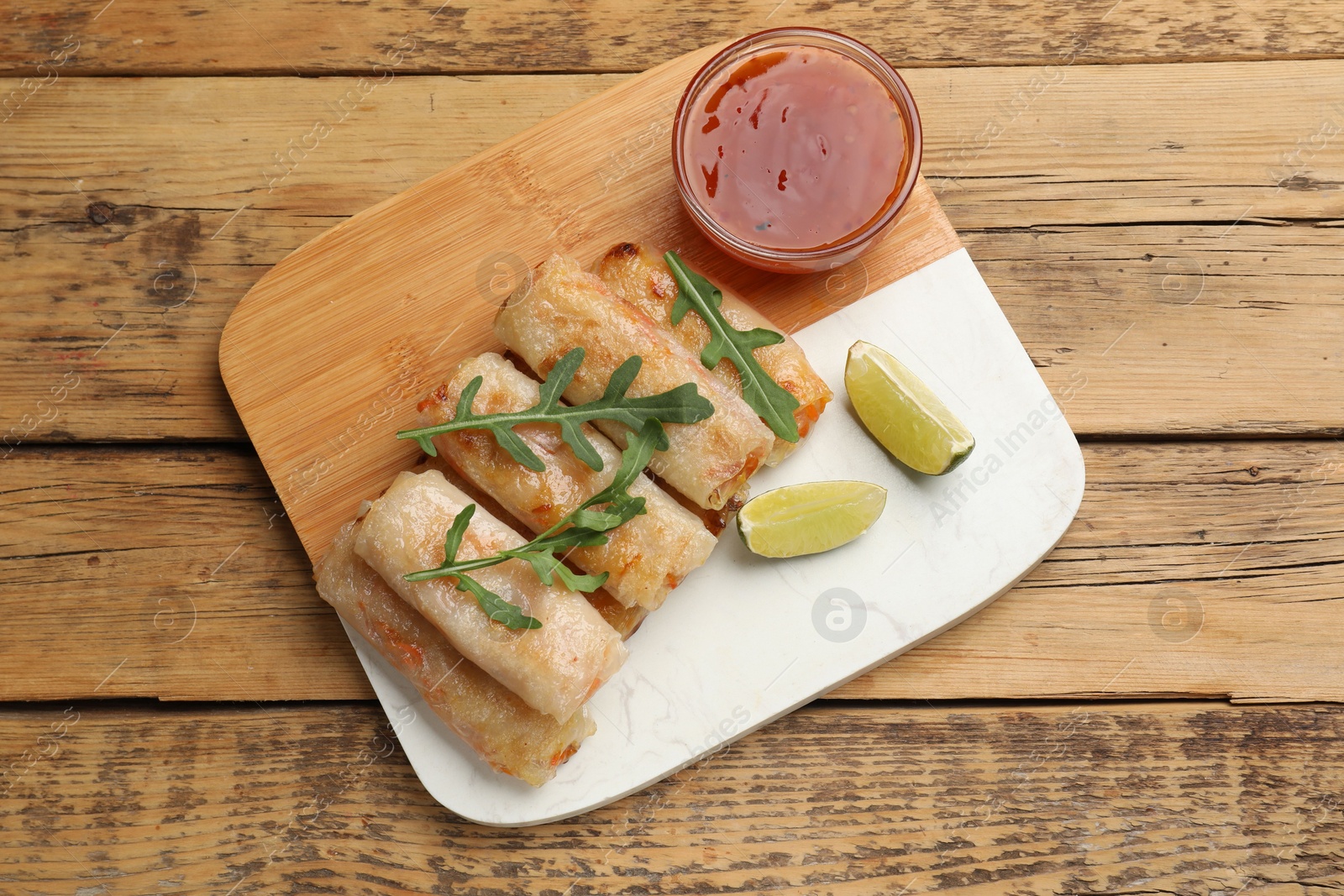 Photo of Tasty fried spring rolls, lime, arugula and sauce on wooden table, top view