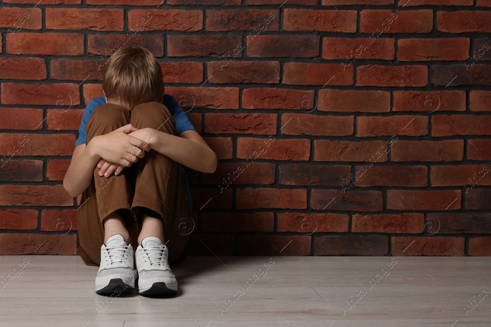 Photo of Upset boy sitting on floor near brick wall, space for text. Children's bullying