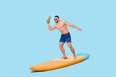 Happy man with pineapple posing on SUP board against light blue background