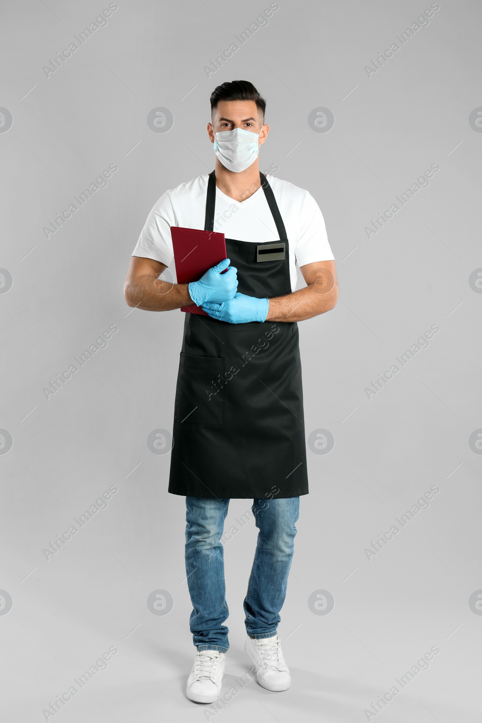 Photo of Waiter in medical face mask with menu on light grey background