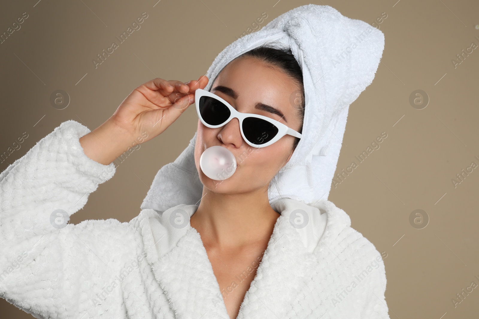 Photo of Young woman in bathrobe and sunglasses blowing chewing gum on brown background