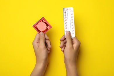 Photo of Woman holding condom and birth control pills on yellow background, top view. Safe sex