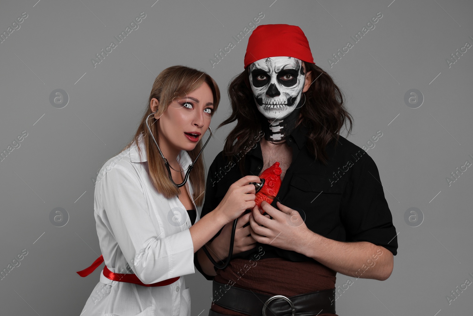 Photo of Couple in scary nurse and pirate costumes with heart model and stethoscope on light grey background. Halloween celebration