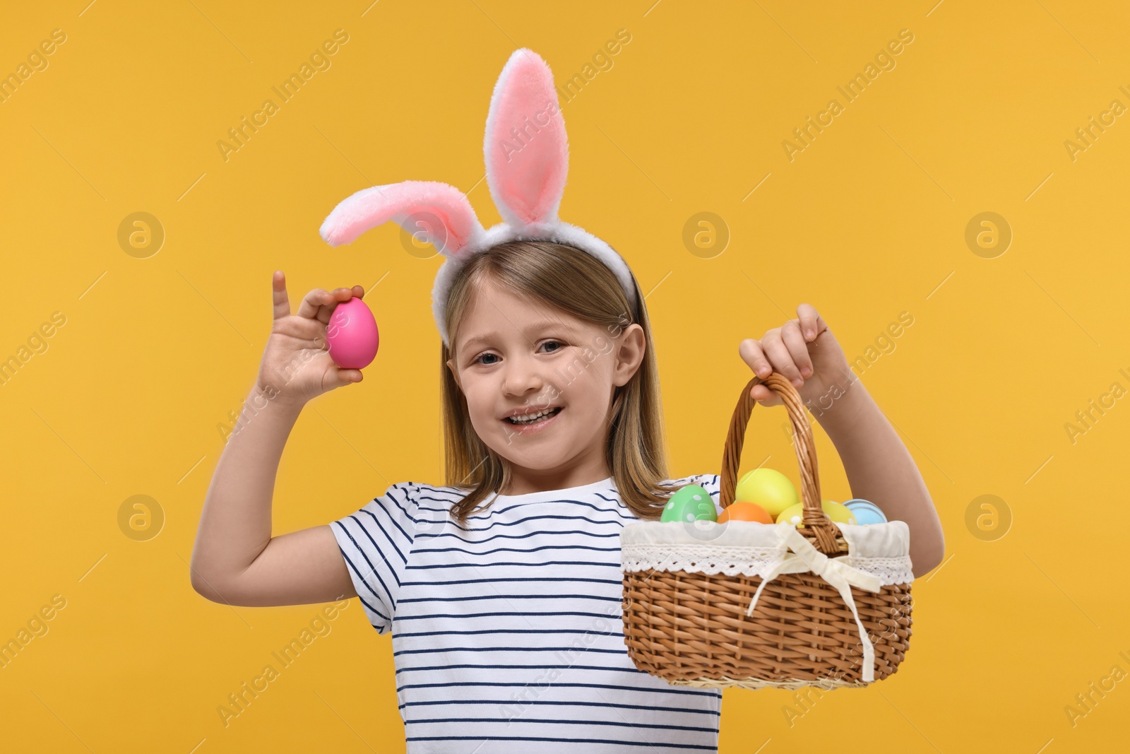 Photo of Easter celebration. Cute girl with bunny ears holding basket of painted eggs on orange background