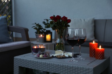 Glasses of wine, vase with roses, burning candles and snacks on rattan table at balcony in evening