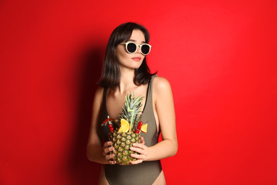 Beautiful woman in stylish swimsuit holding tropical cocktail on red background