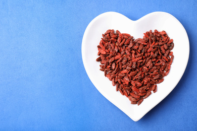 Photo of Dried goji berries in heart shaped plate on blue background, top view. Space for text