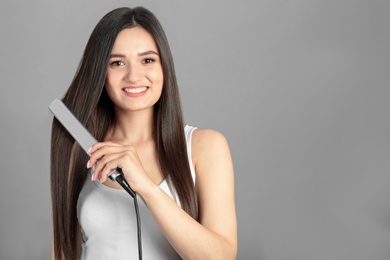 Young woman using hair iron on grey background, space for text