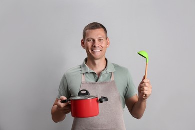 Happy man with cooking pot and ladle on light grey background