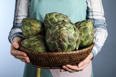 Photo of Woman holding wicker basket with fresh raw artichokes on grey background, closeup