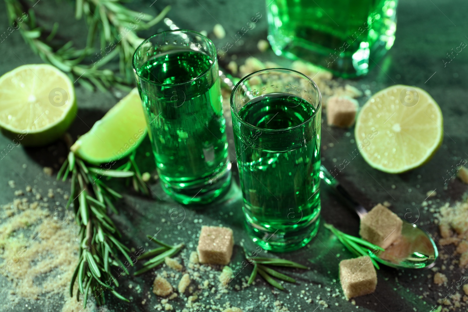 Photo of Absinthe in shot glasses, spoon, brown sugar, lime and rosemary on gray table, closeup. Alcoholic drink