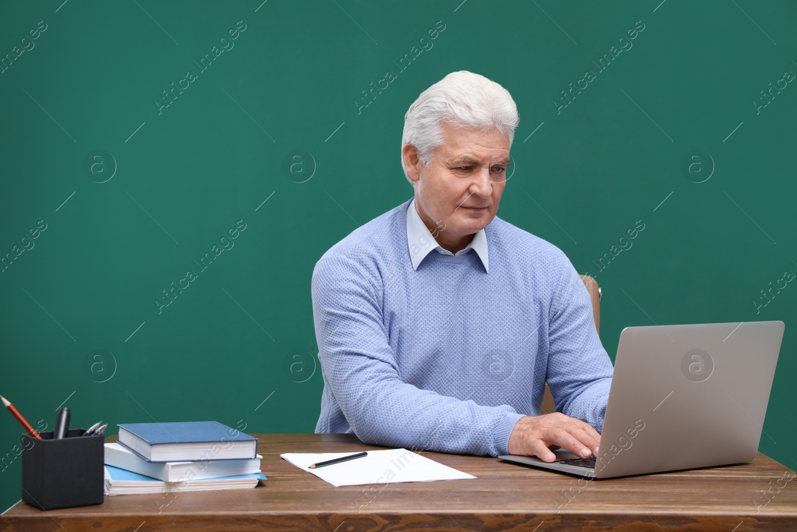 Photo of Portrait of senior teacher with laptop at table against green chalkboard