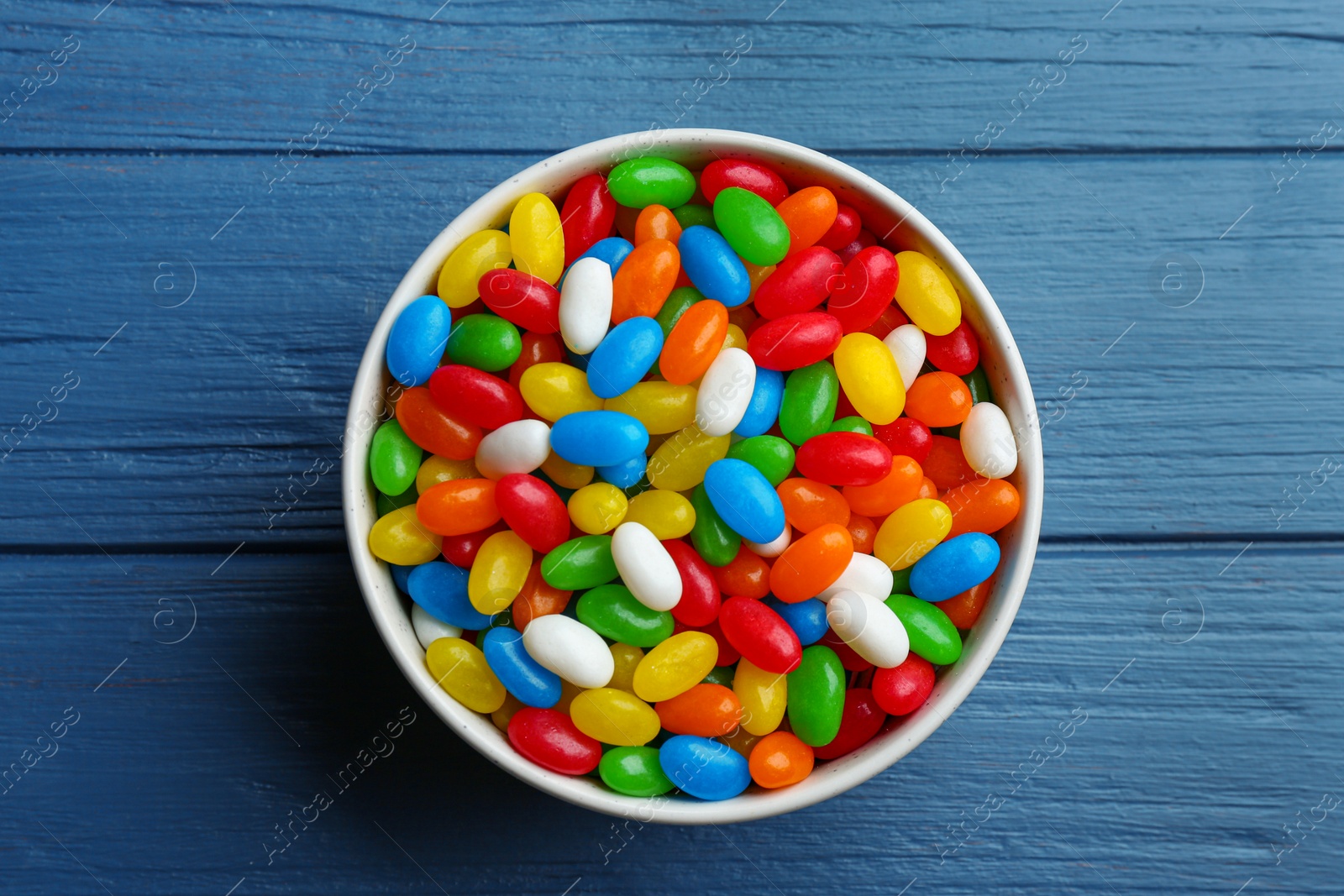 Photo of Bowl with colorful jelly beans on blue wooden background, top view