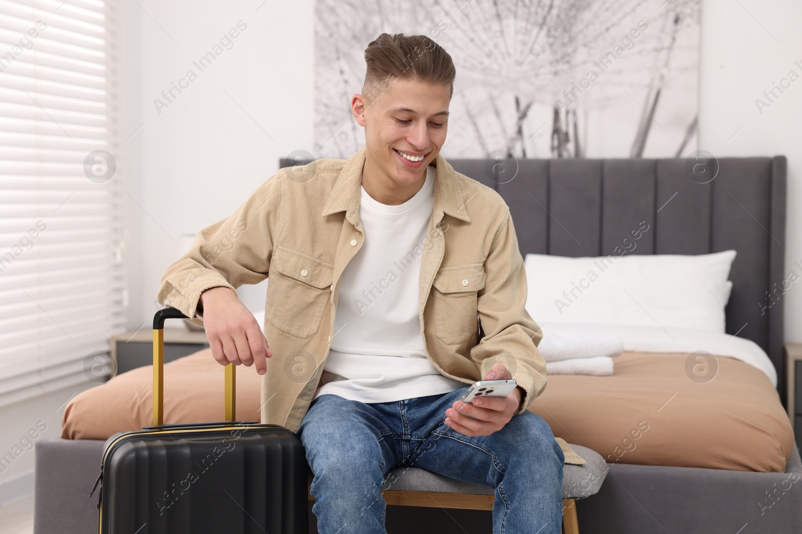 Photo of Smiling guest with smartphone and suitcase in stylish hotel room