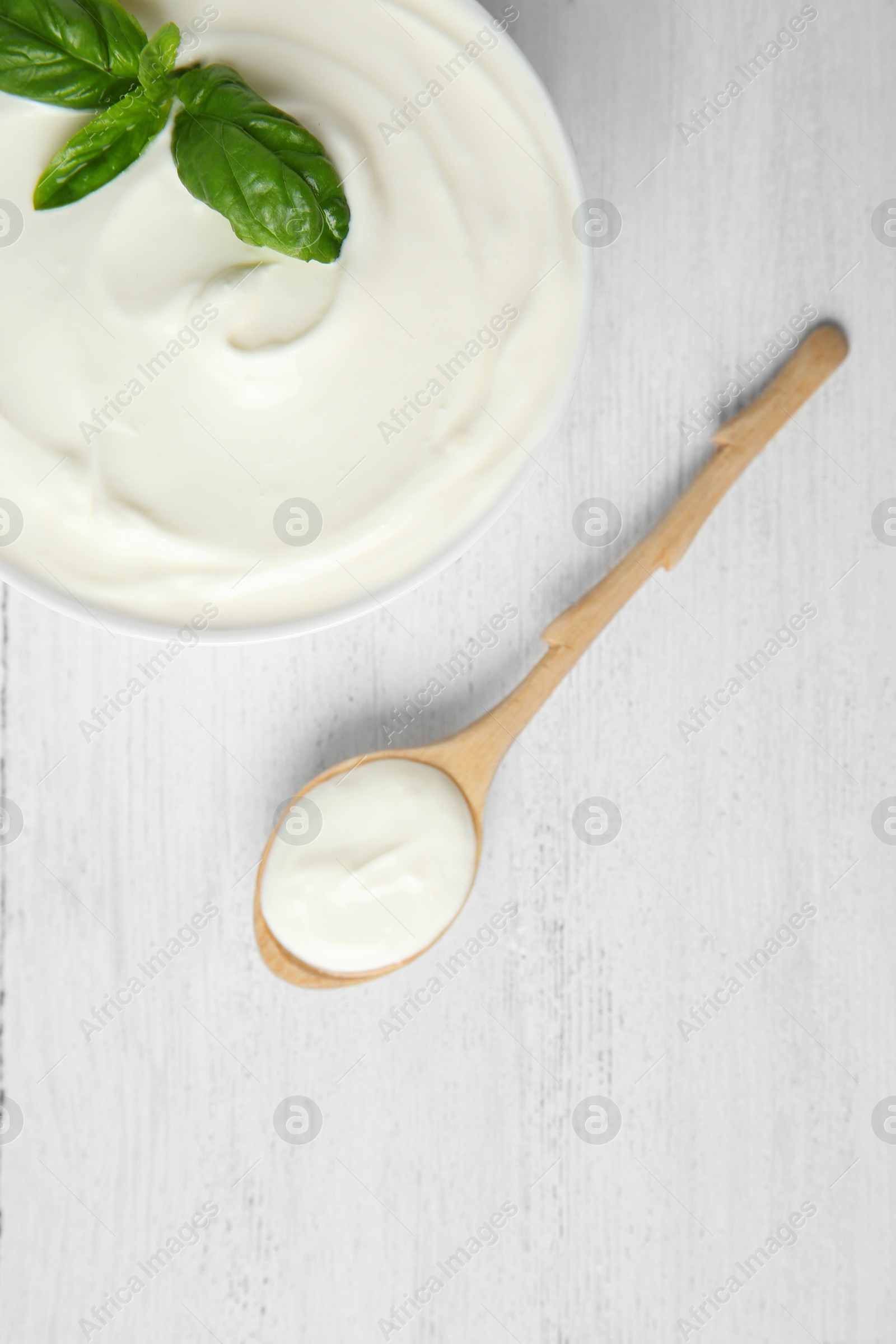 Photo of Bowl of fresh sour cream with basil and spoon on white wooden table, flat lay