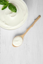 Photo of Bowl of fresh sour cream with basil and spoon on white wooden table, flat lay