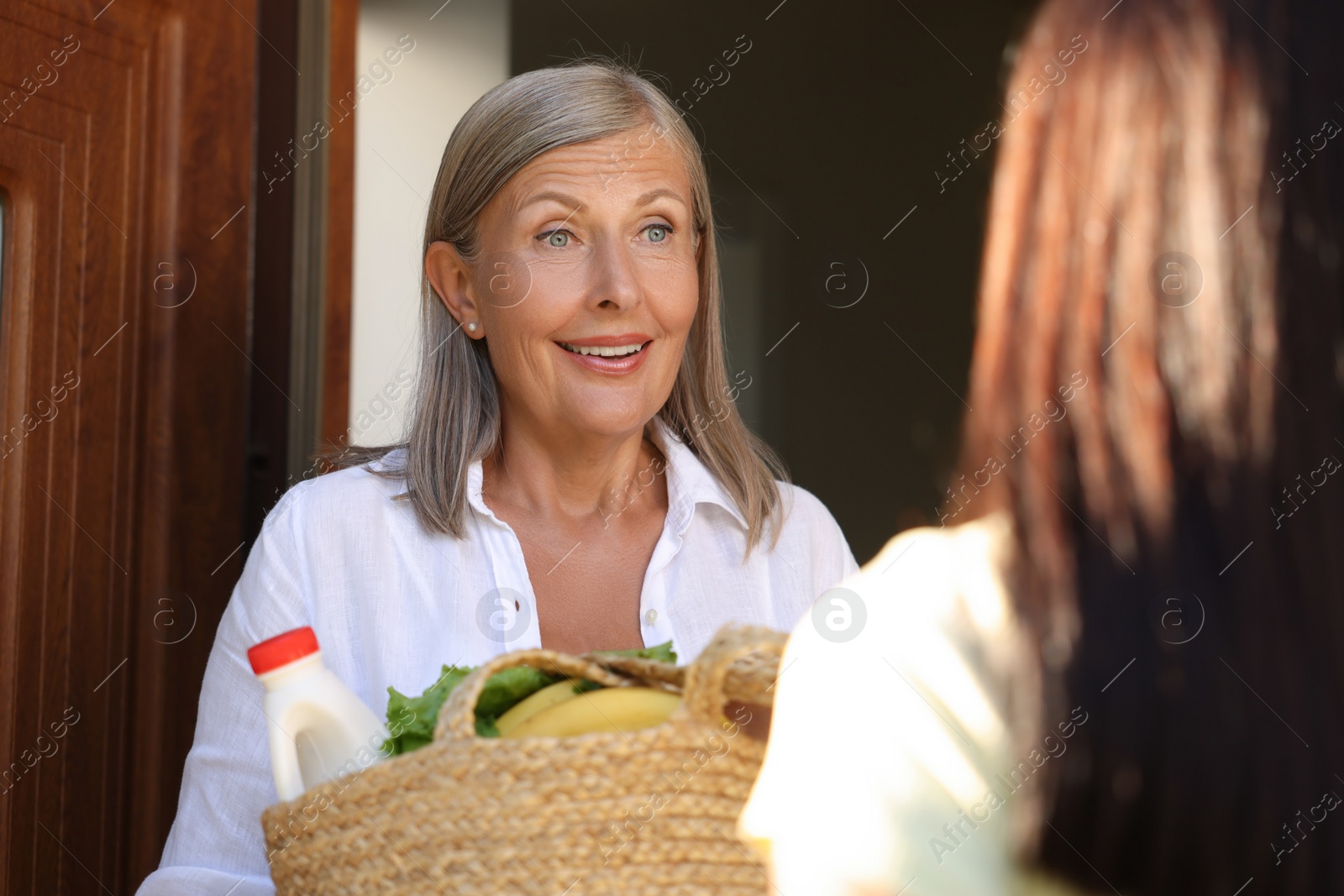 Photo of Young woman with wicker bag of products helping her senior neighbour outdoors