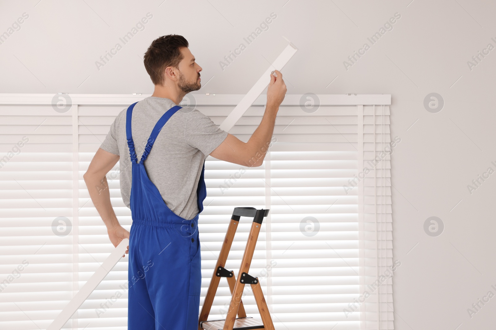 Photo of Worker in uniform installing horizontal window blinds on stepladder indoors