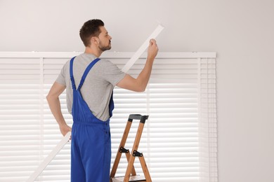 Worker in uniform installing horizontal window blinds on stepladder indoors