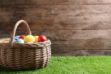 Photo of Wicker basket with painted Easter eggs on green grass against wooden background, space for text