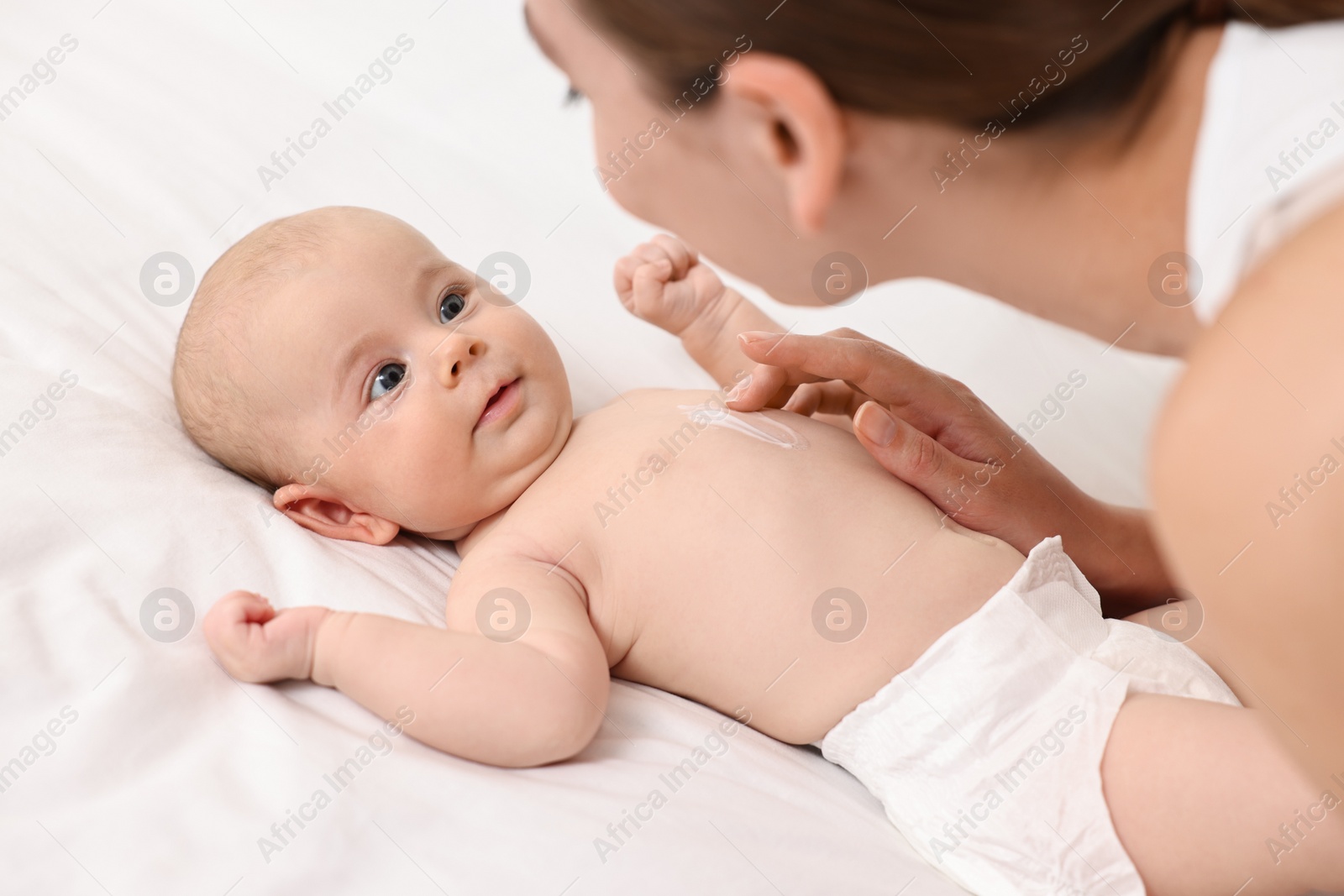 Photo of Woman applying body cream onto baby`s skin on bed, closeup