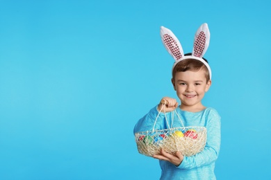 Photo of Little boy in bunny ears headband holding basket with Easter eggs on color background, space for text