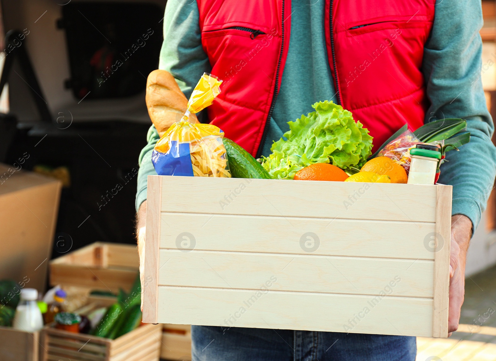Photo of Courier holding crate with products near car outdoors, closeup. Food delivery service