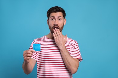 Emotional man holding condom on light blue background. Safe sex