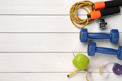 Dumbbells, skipping rope, apple and measuring tape on white wooden table, flat lay. Space for text