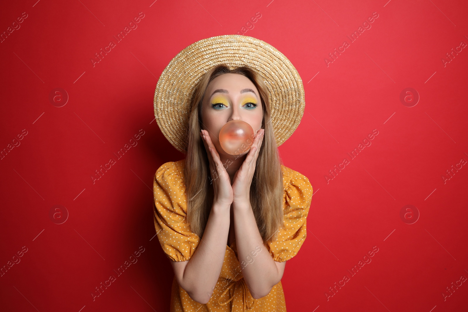 Photo of Fashionable young woman with bright makeup blowing bubblegum on red background