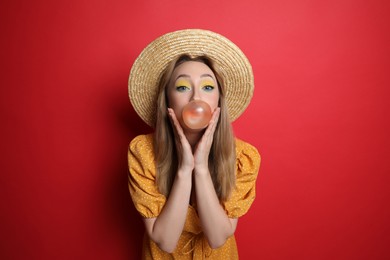 Fashionable young woman with bright makeup blowing bubblegum on red background