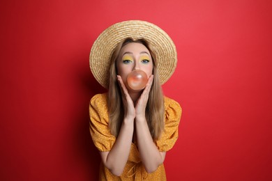 Photo of Fashionable young woman with bright makeup blowing bubblegum on red background