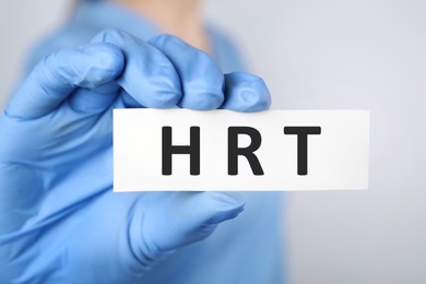 Image of Doctor holding card with abbreviation HRT on light grey background, closeup. Hormone Replacement Therapy