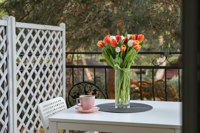 Photo of Beautiful colorful tulips in glass vase and cup of drink on white table at balcony