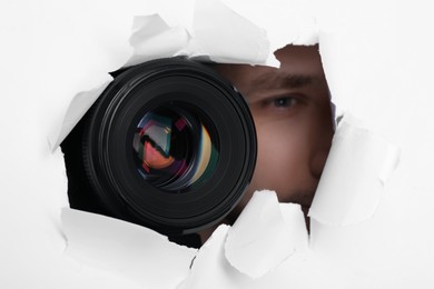 Hidden man with camera spying through torn hole in white paper, closeup