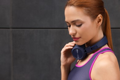 Beautiful woman in stylish gym clothes with headphones posing near dark grey wall on street, closeup. Space for text