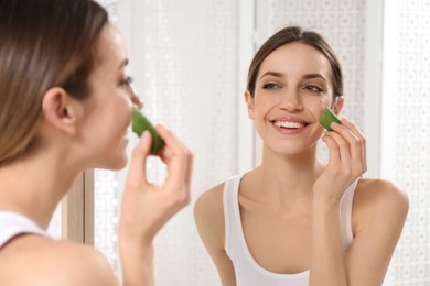 Photo of Young woman using aloe leaf to take care of skin near mirror indoors