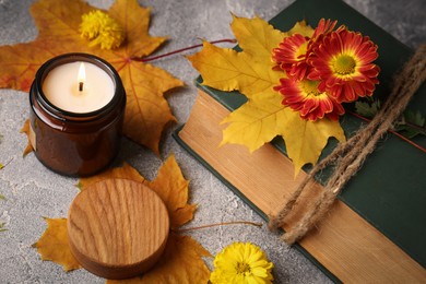 Burning scented candle, autumn leaves and book with beautiful flowers on light gray textured table