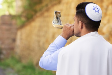 Photo of Jewish man in kippah and tallit blowing shofar outdoors. Rosh Hashanah celebration