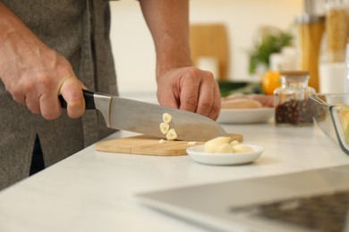 Photo of Man cutting garlic while watching online cooking course via laptop in kitchen, closeup