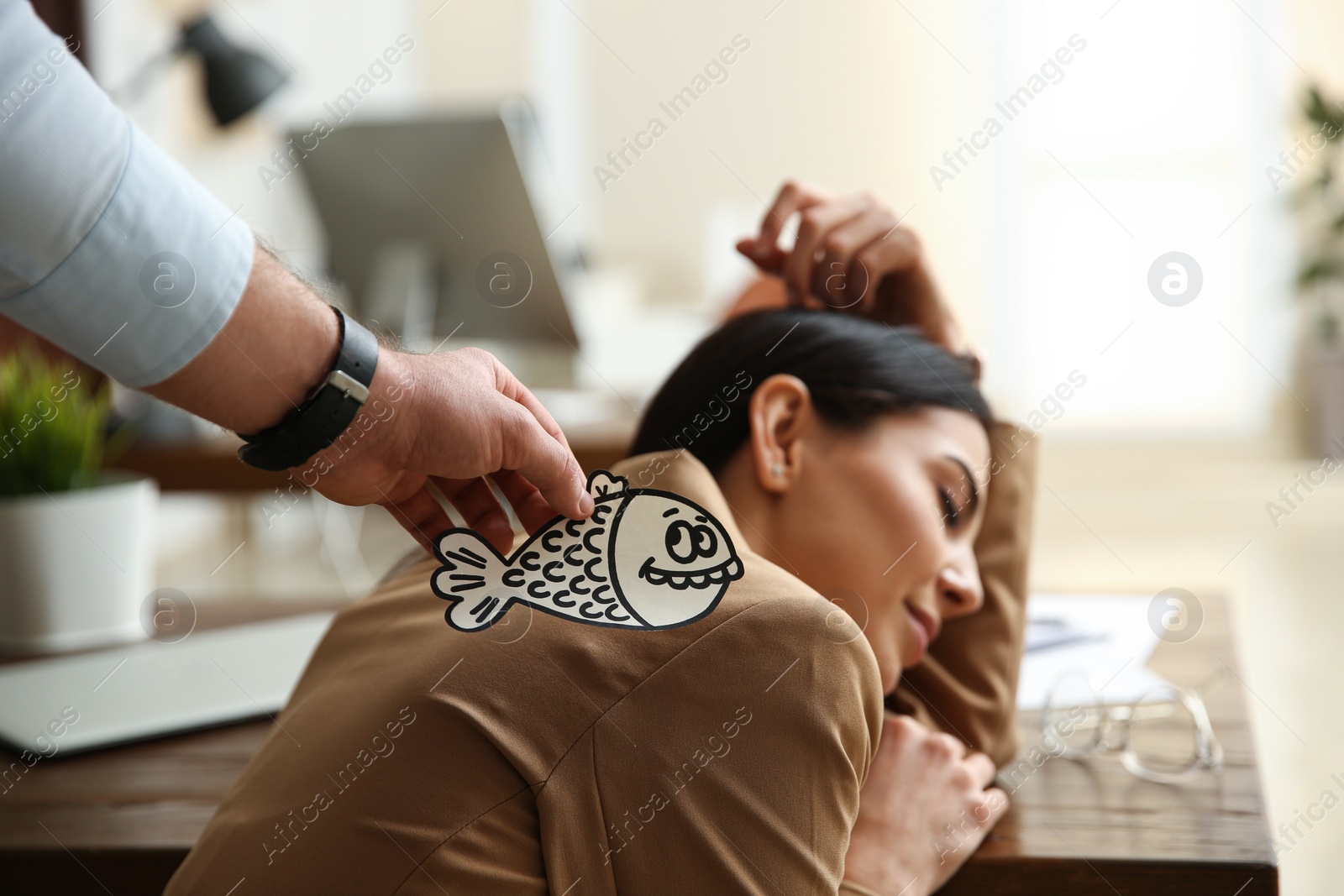 Photo of Man sticking paper fish to colleague's back while she sleeping in office, closeup. Funny joke