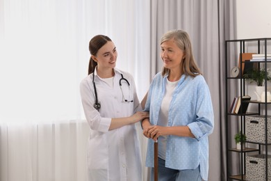 Photo of Young healthcare worker assisting senior woman with walking cane indoors