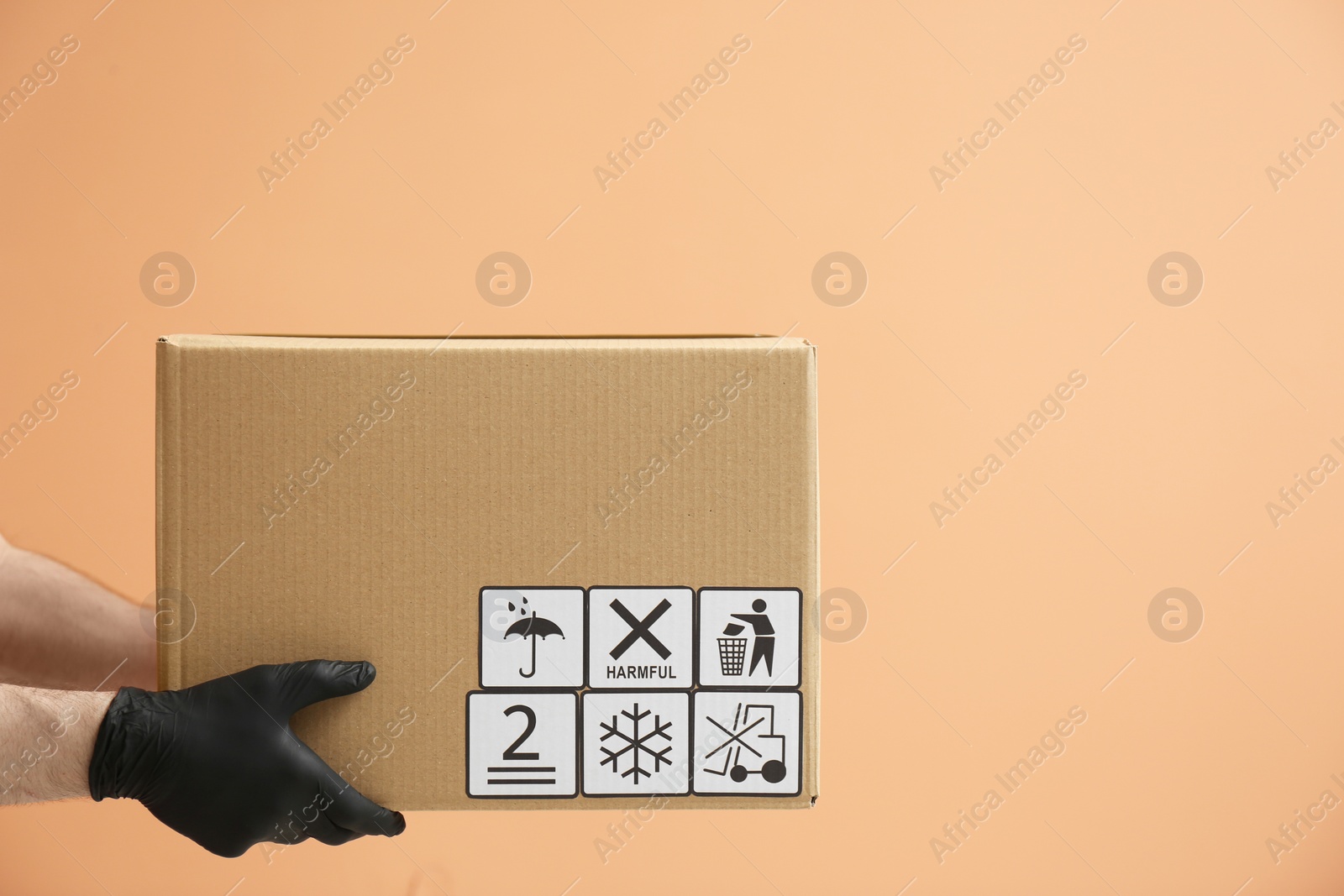 Photo of Courier holding cardboard box with different packaging symbols on orange background, closeup. Parcel delivery