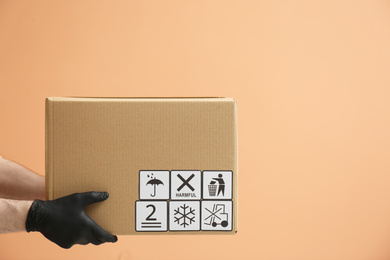 Photo of Courier holding cardboard box with different packaging symbols on orange background, closeup. Parcel delivery